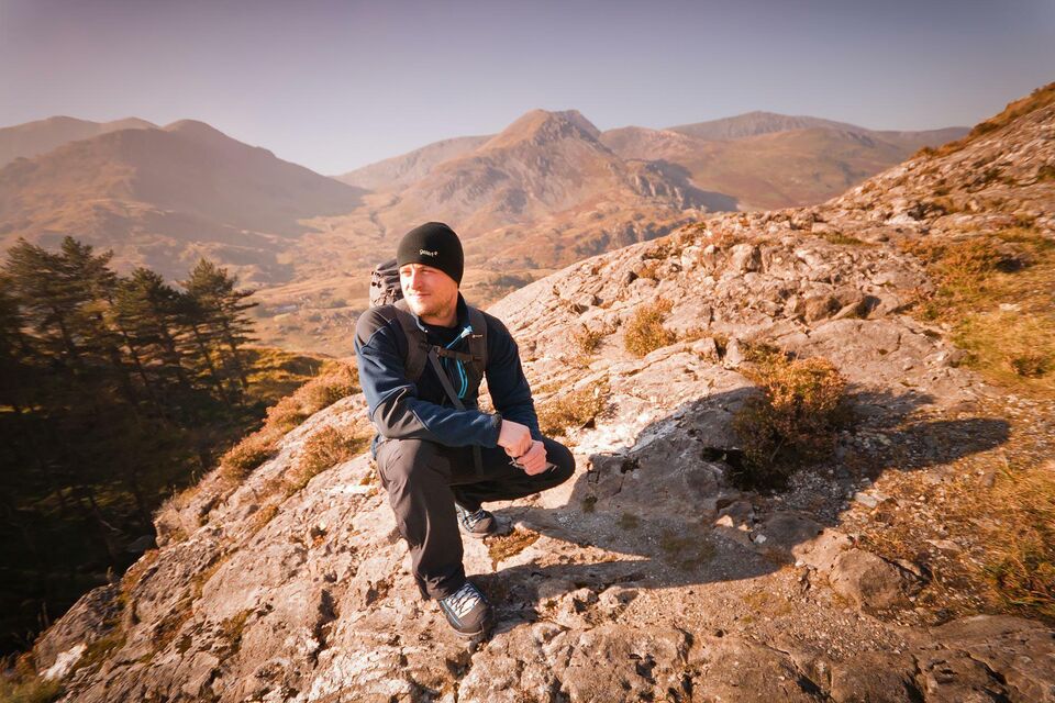 A man in winter walking gear crouching on a rocky outcrop in the Welsh mountains.